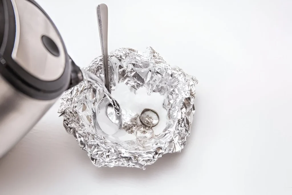 Water is being poured into silver silverware in baking soda and salt-soaked solution in a dish covered with aluminium foil